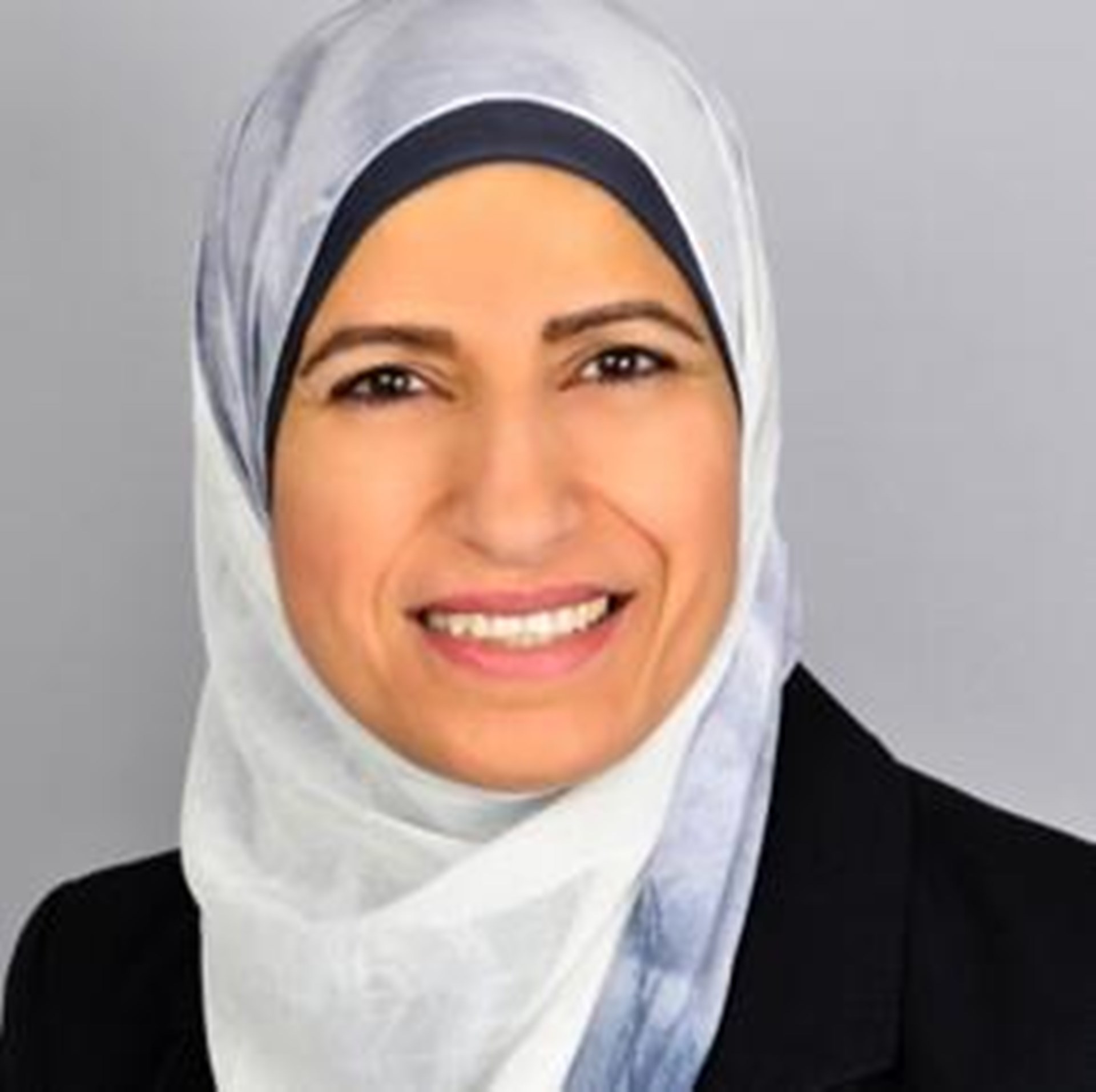 Frau mit grauem Kopftuch und schwarzem Blazer lächelt vor grauem Hintergrund.