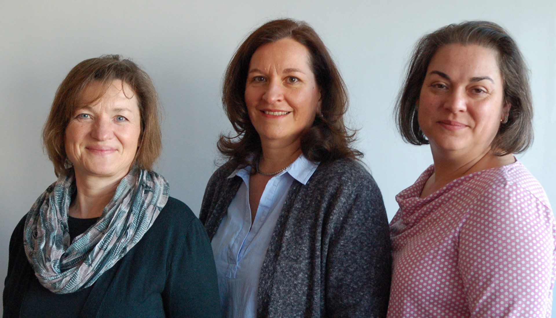 Drei lächelnde Frauen in schicker Kleidung stehen vor hellem Hintergrund.