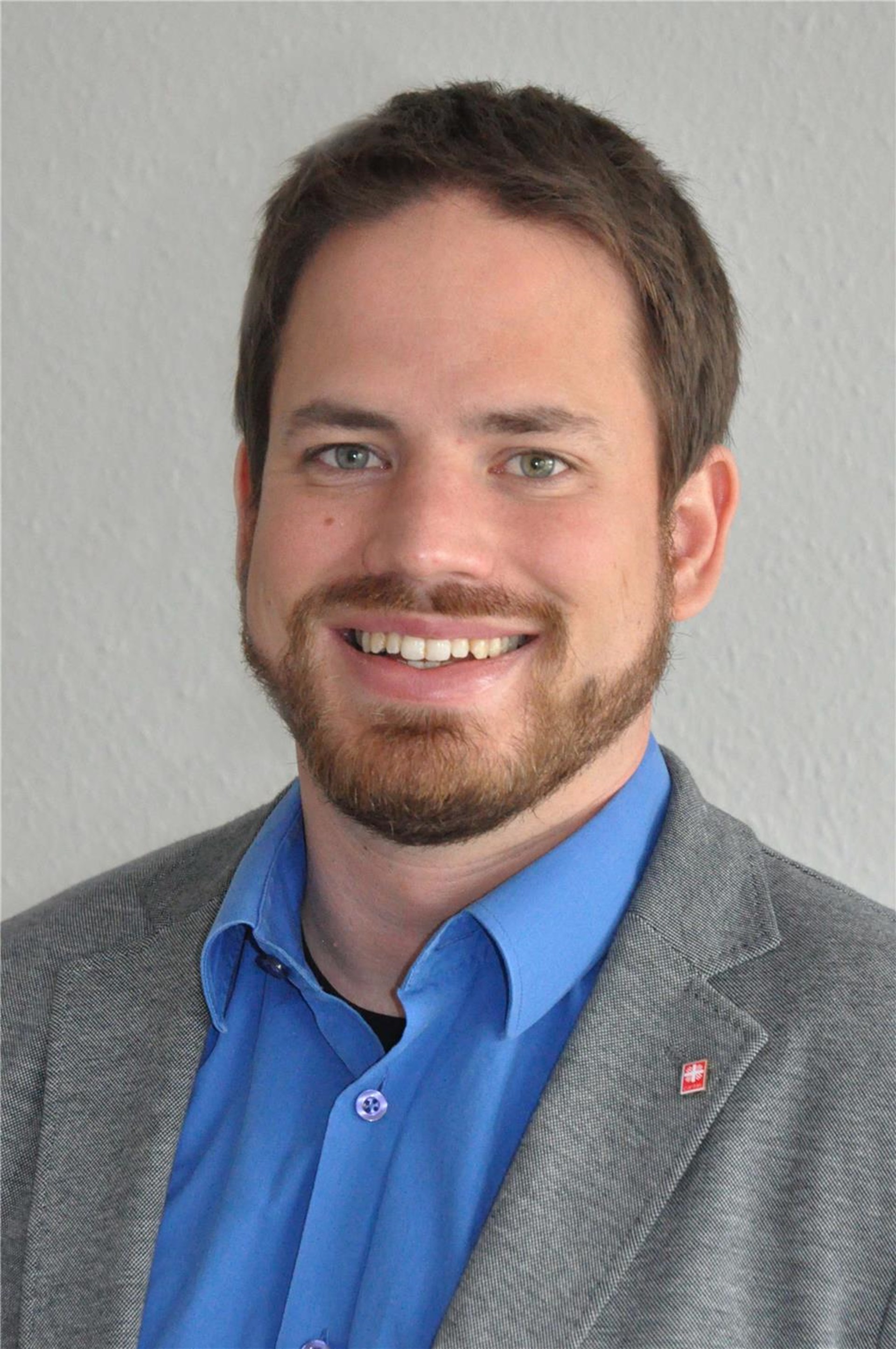 Lächelnder Mann mit braunem Bart, blauem Hemd und grauem Sakko vor hellem Hintergrund.