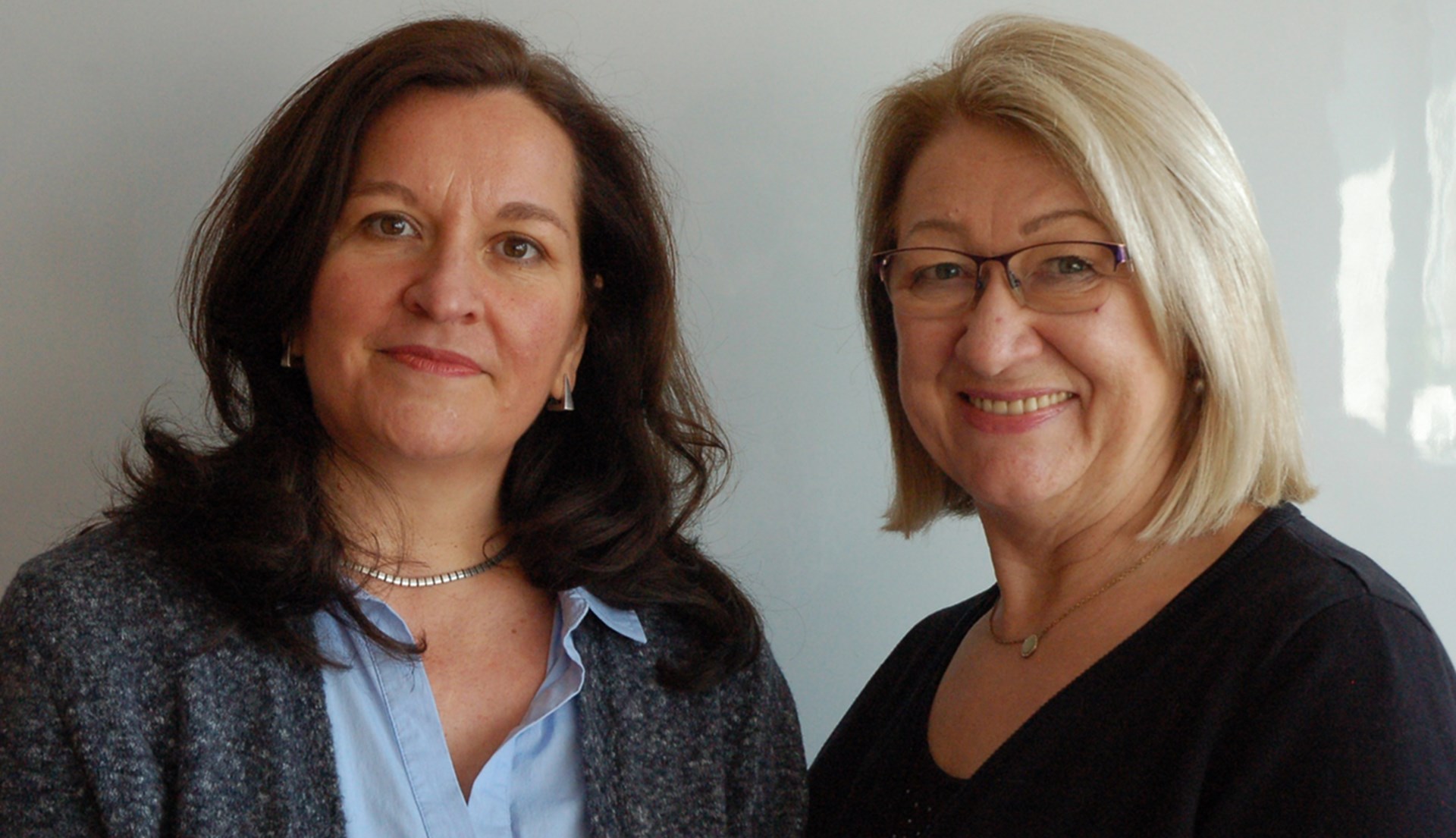 Zwei lächelnde Frauen, eine mit braunen, eine mit blonden Haaren, vor hellem Hintergrund.