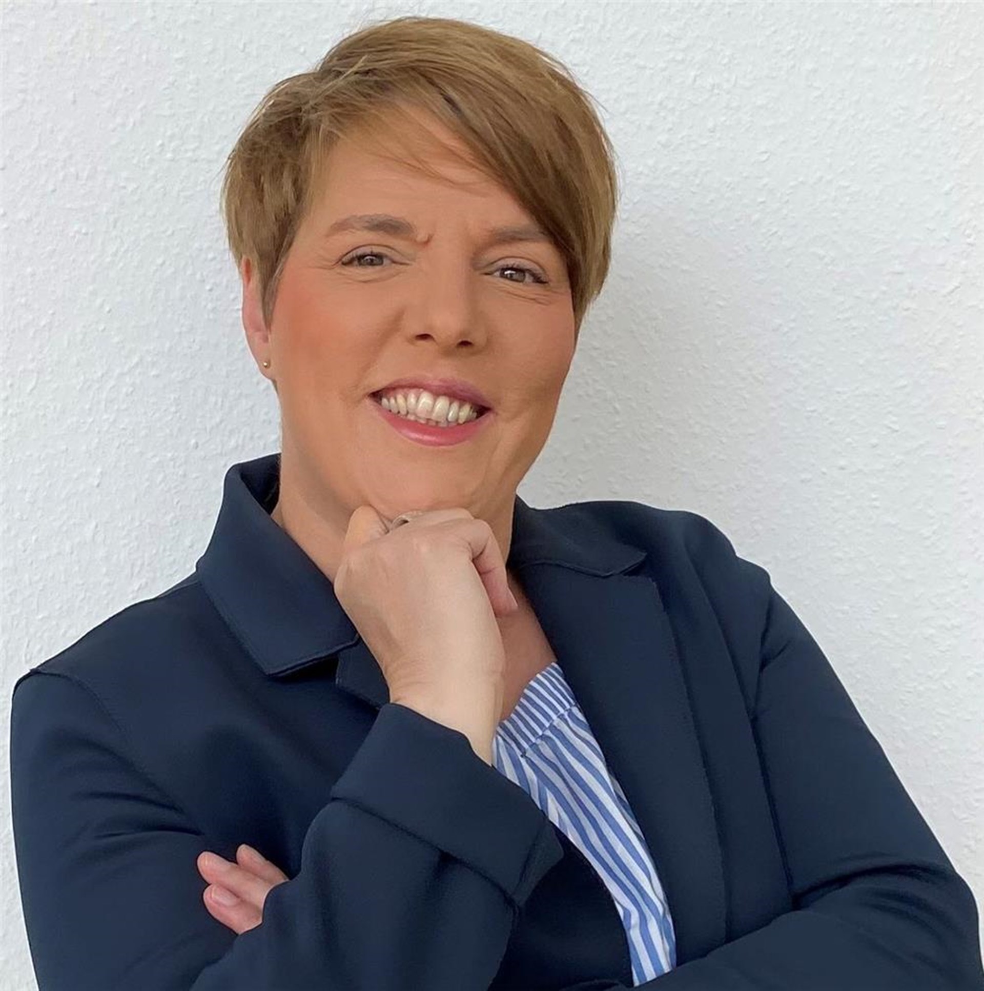 Porträt einer Frau mit kurzem Haar in dunkelblauem Blazer, lächelnd mit Hand am Kinn.