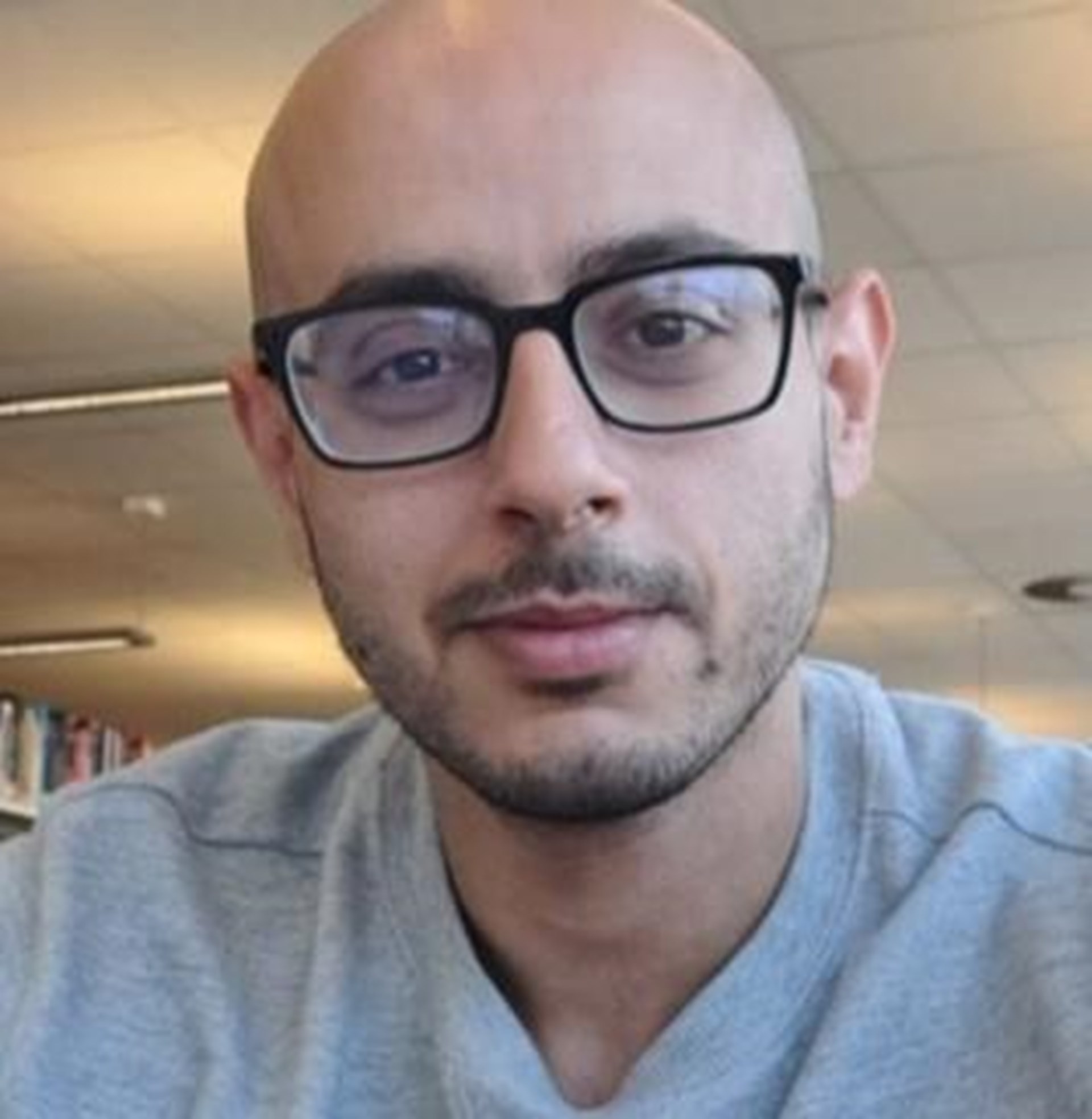 Mann mit Brille und grauem Pullover sitzt in einem hellen Raum mit Bücherregal.